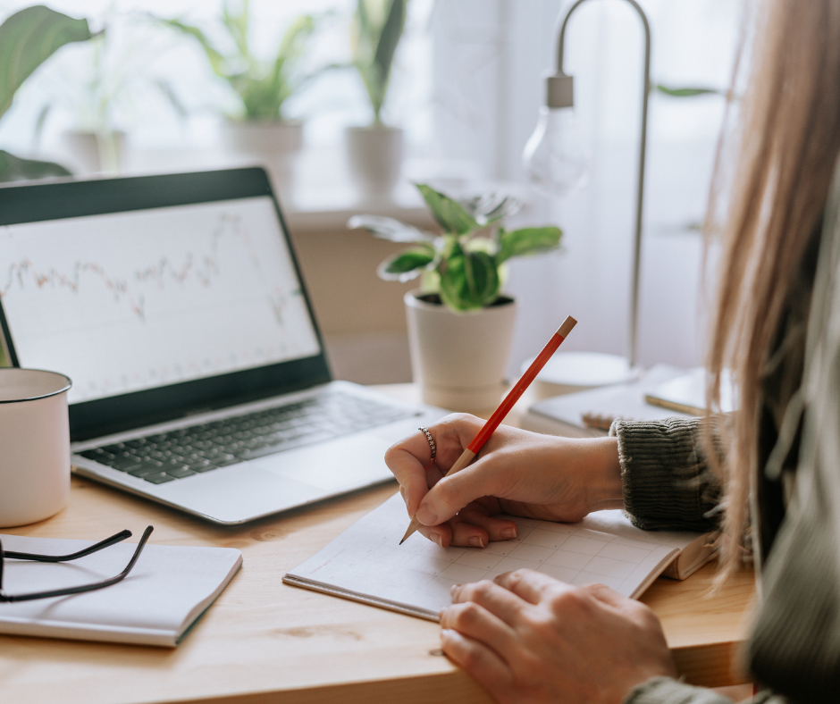 Image of someone sitting at a desk writing in a notepad.  There's also an open laptop, a mug and several pot plants dotted around